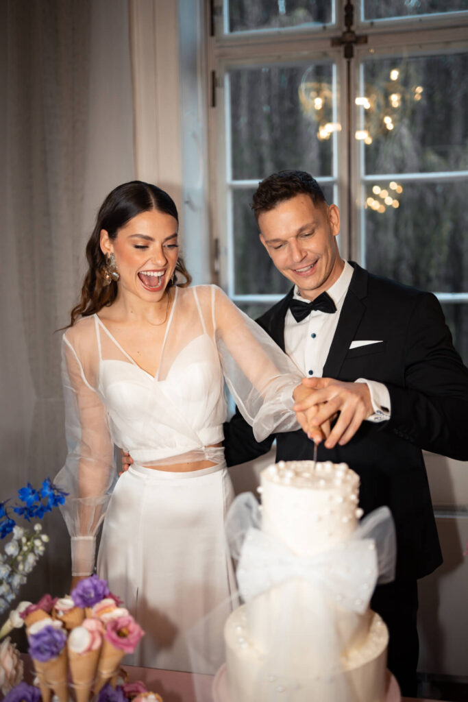 modernes Brautpaar schneidet gemeinsam Hochzeitstorte an und lacht dabei ausgelassen.