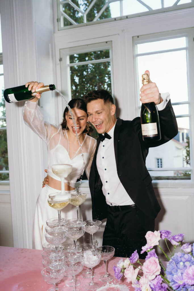 Brautpaar schüttet Champagner in Gläser ein und lacht ausgetobt.