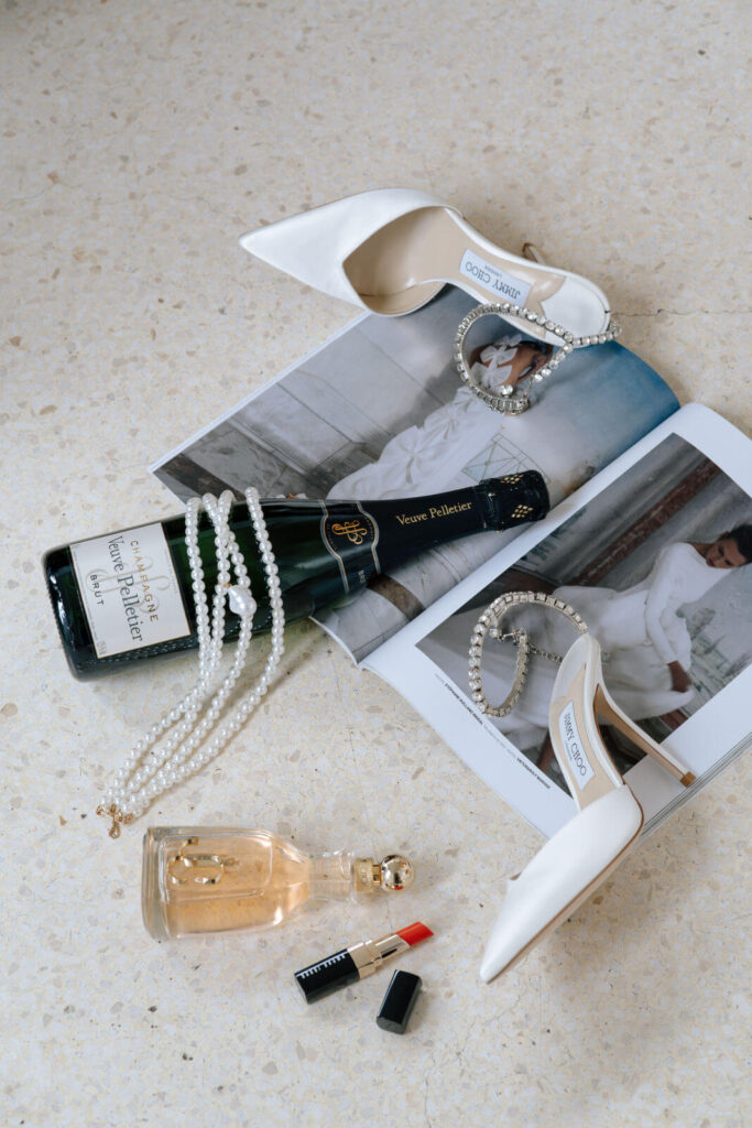 Schuhe der Braut liegt mit Lippenstift und Champagner und Perlenkette vor einem Brautmagazin auf dem Boden.
