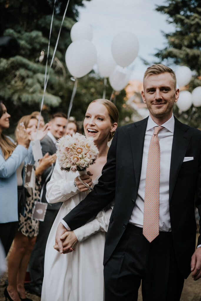 Brautpaar hält sich an den Händen und geht an Gäste vorbei, Braut hält Blumenstrauß in der Hand und weiße Luftballons fliegen im Hintergrund.