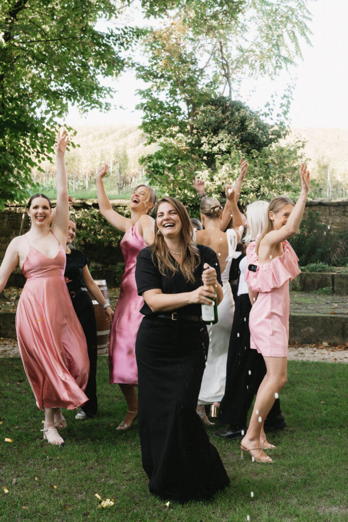 Braut steht mit Brautjungfern im Garten, Braut sprüht Champagnerflasche, Frauen tanzen im Hintergrund.