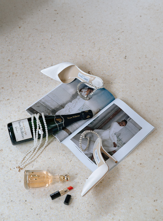 Champagnerflasche, weiße Dior Schuhe und Perlenkette liegen auf Hochzeitsmagazin am Boden.