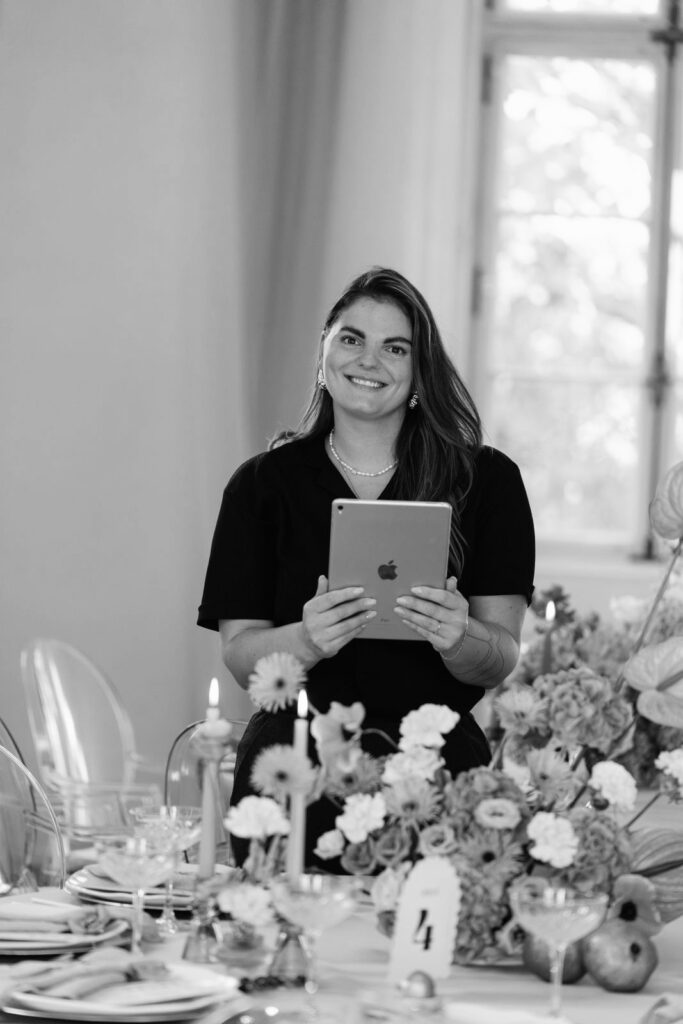 Hochzeitsplanerin Lisa steht mit ihrem iPad vor einem Tisch mit Blumen und lächelt in die Kamera